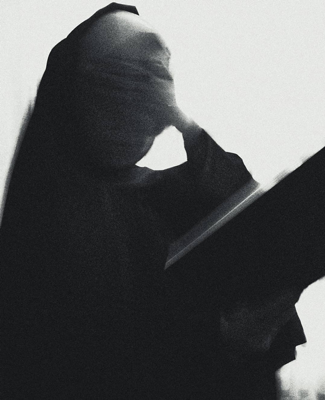 Black and white photograph of a nun covering her face with one hand and an open bible in the other hand.