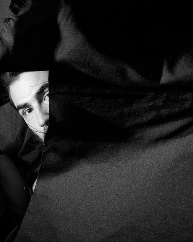 Black and white photograph of a man looking at the camera behind some fabrics that are in the air.