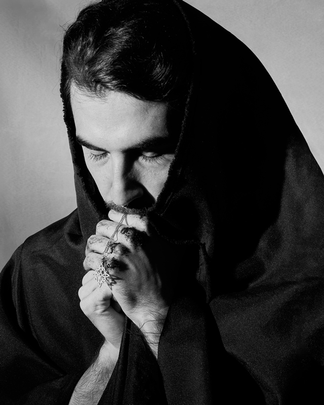 Black and white photograph of a man with a tunic on his head, praying with his hands on his face.