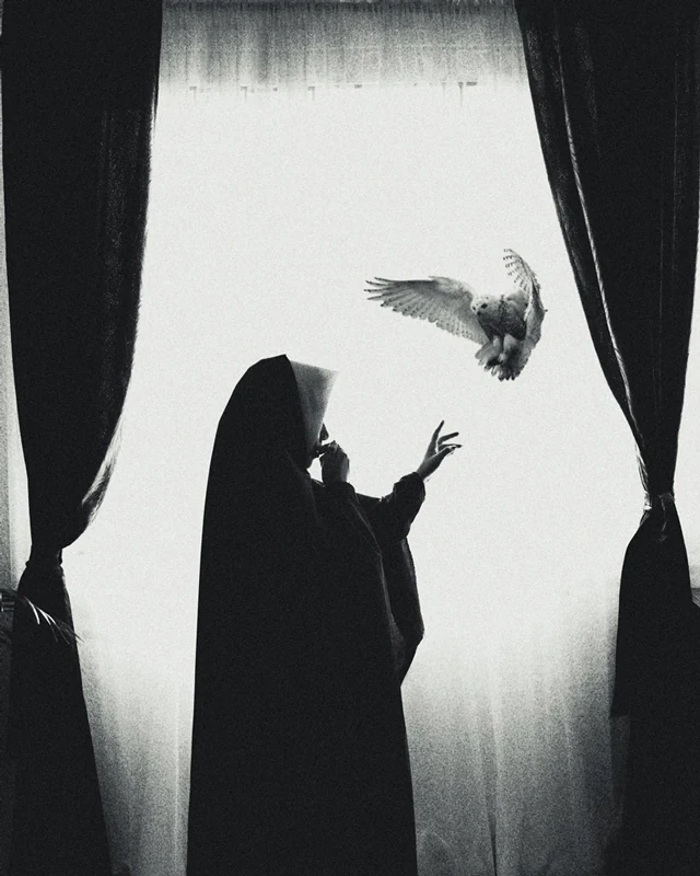Black and white photograph nun in front of a giant window, holding an owl in her hand.