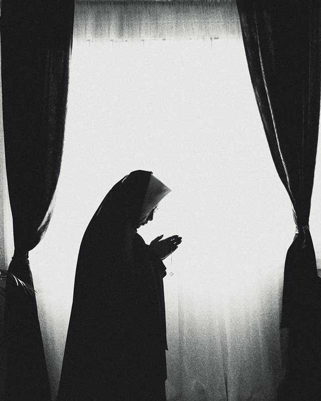 Black and white photograph nun in front of a giant window praying.