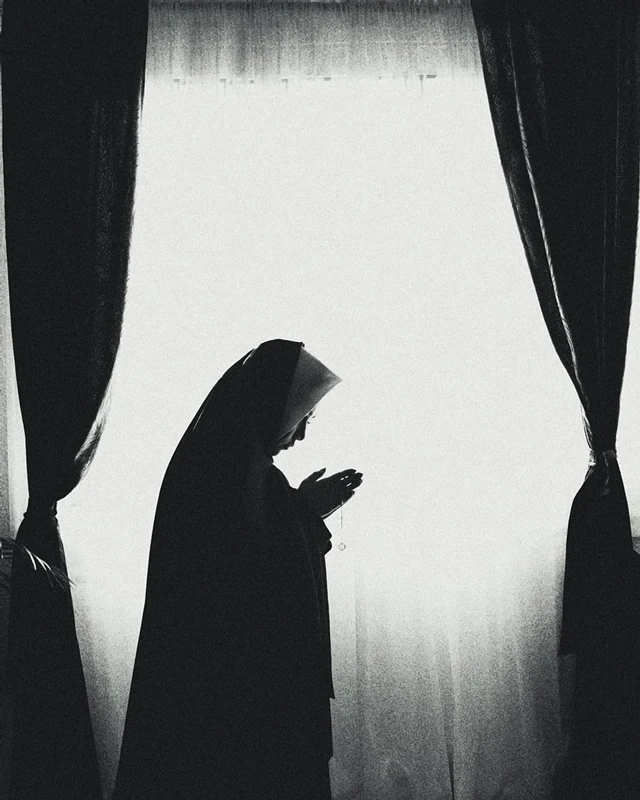 Black and white photograph nun in front of a giant window praying.