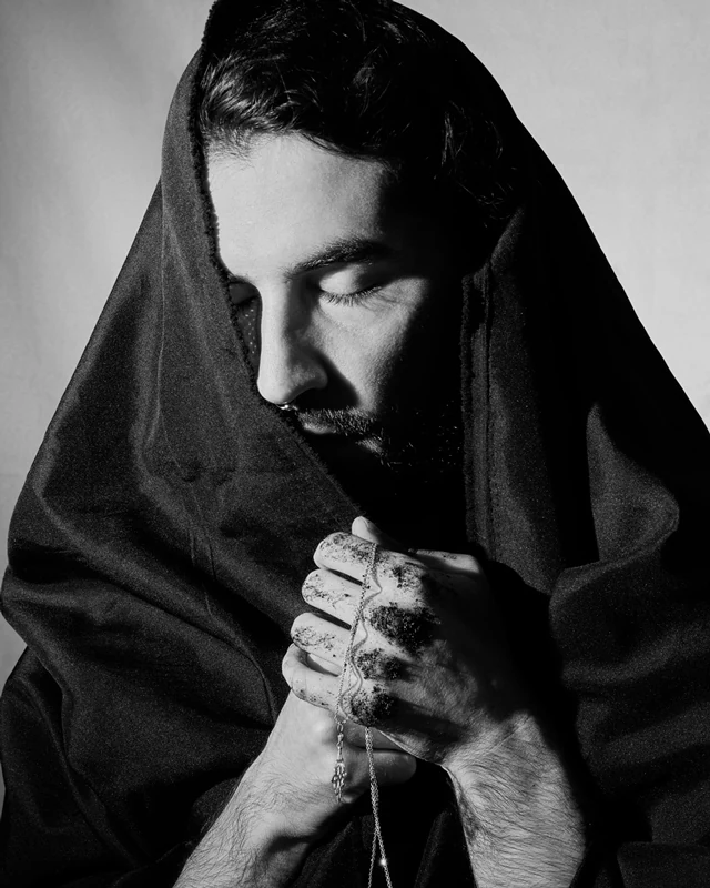 Black and white portrait photograph of a man wearing a tunic on his head, with his eyes closed and a chain in his hands.