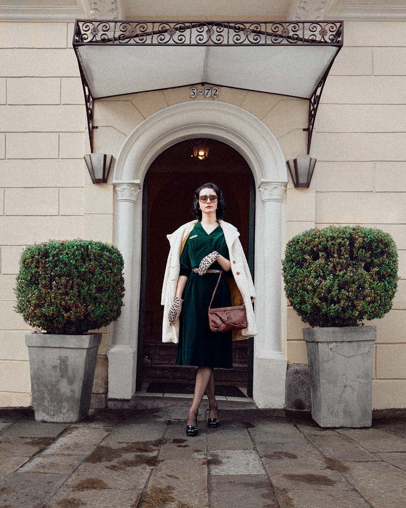 Portada editorial mujer elegante con saco en sostenido en los hombros, vestido verde y una cartera de color café, en la puerta de un hotel