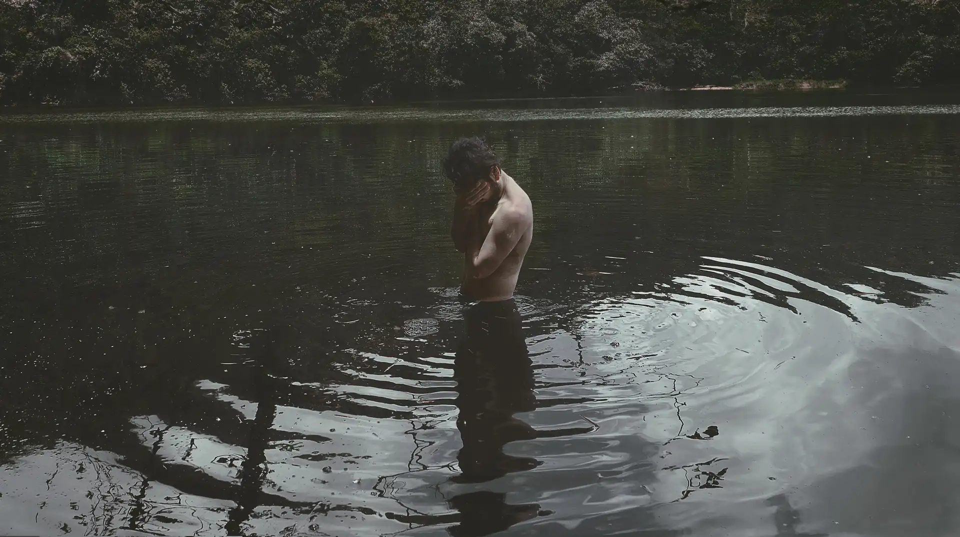Hombre con medio cuerpo sumergido en un lago, con las manos tapando su rostro