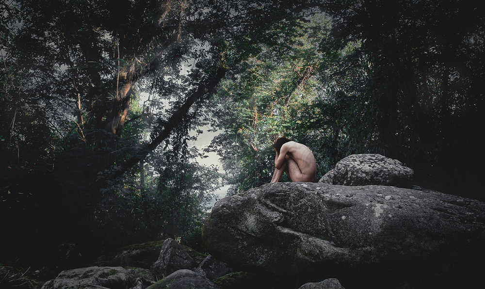 Artistic photograph of a man in a forest, sitting on a giant stone covering his face against his knees.