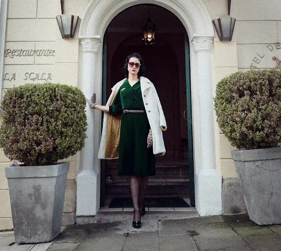 Photograph of an elegant woman dressed in an emerald green dress and gloves, leaving a hotel.