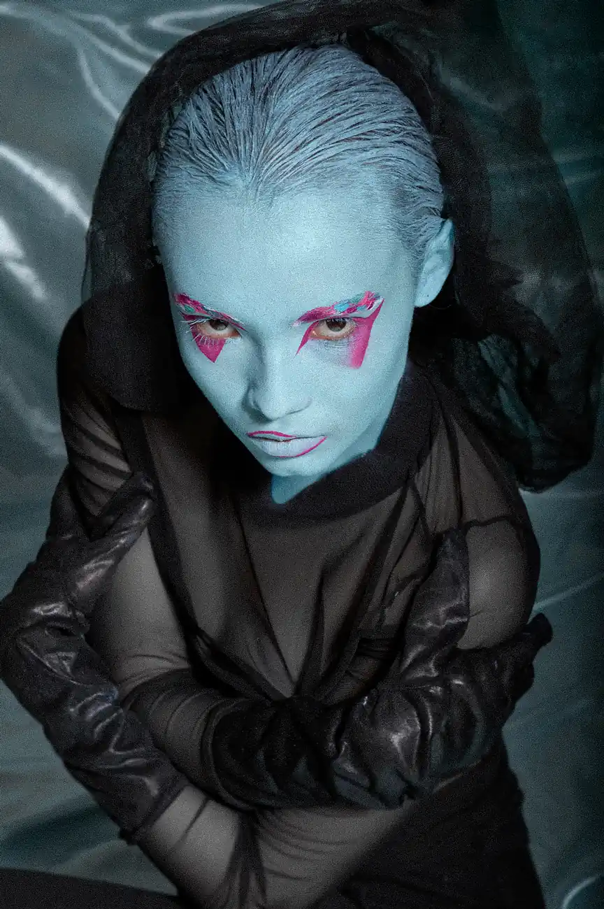 Mujer con maquillaje azul y sombras rosadas, con los brazos cruzados