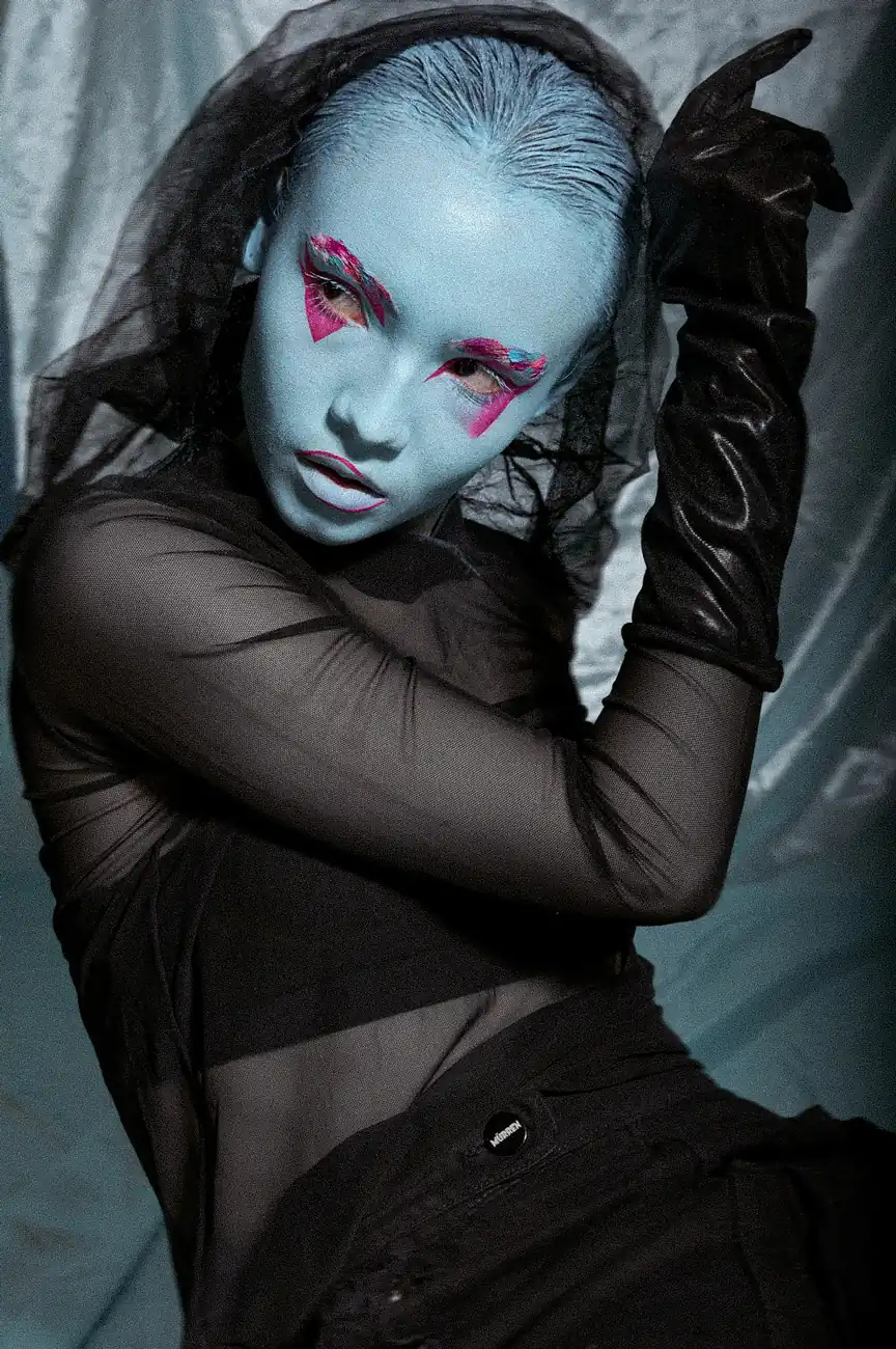 Mujer con maquillaje azul y sombras rosadas, mirando el horizonte con la mano en el rostro