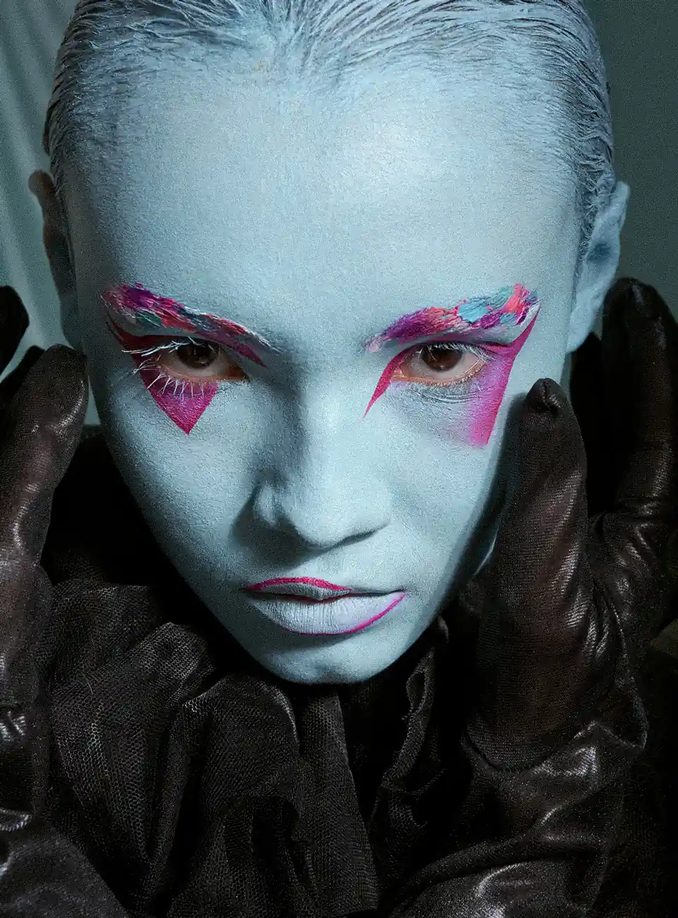 Mujer con maquillaje azul y sombras rosadas, con sus dos manos al rededor del rostro