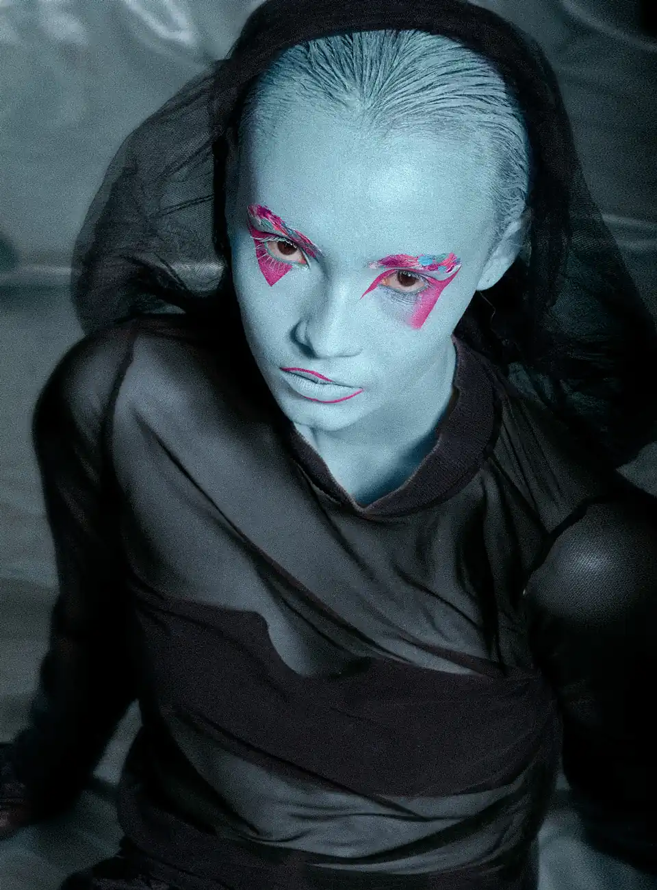 Mujer con maquillaje azul y sombras rosadas, sentada en el suelo, mirando desde abajo a la cámara